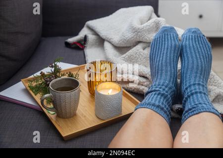 Soft photo of a woman`s legs in woolen blue socks on the sofa with a book and cup of tea and a candle on the tray. Interior and home coziness concept. Stock Photo